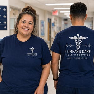 May include: Two people wearing navy blue t-shirts with the Compass Care Health Services logo. The front of the shirt has a small logo, while the back features a larger design with a caduceus symbol, wings, and text. The text reads "Caring. Healing. Helping."