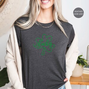 May include: A heather dark grey t-shirt with a green shamrock design and the word "Lucky" written inside the shamrock. The shirt is being modeled by a person.
