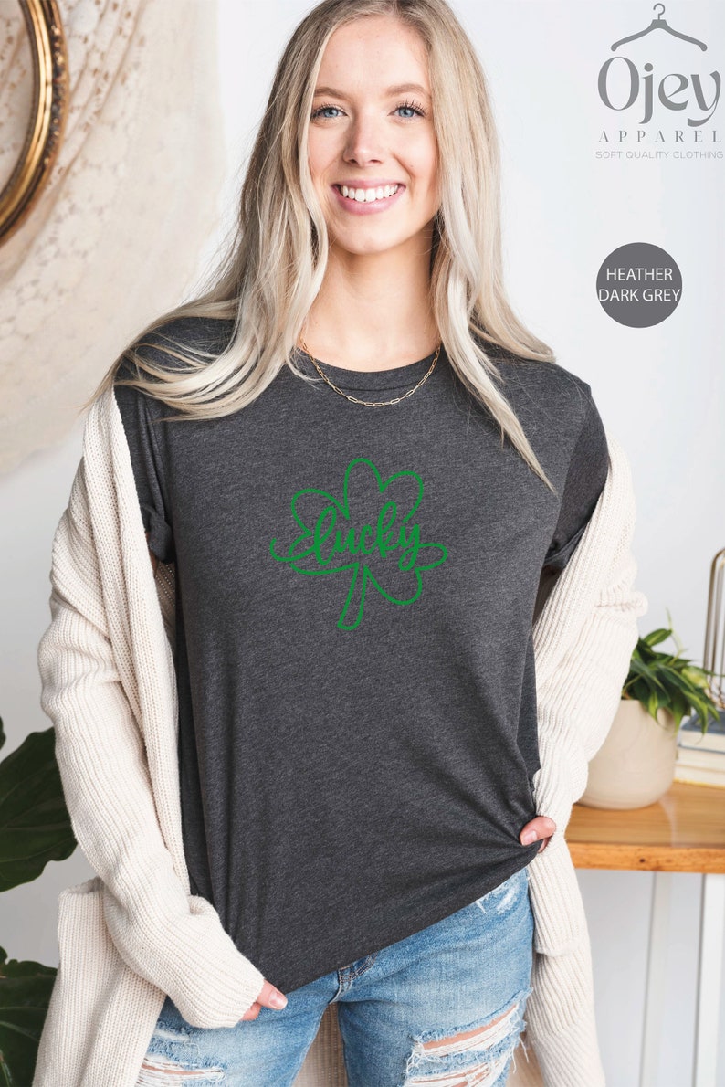May include: A heather dark grey t-shirt with a green shamrock design and the word "Lucky" written inside the shamrock. The shirt is being modeled by a person.