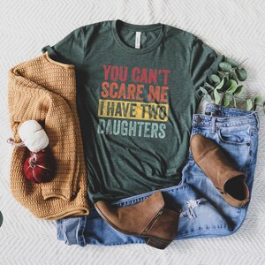 Puede incluir: Una camiseta verde con el texto "You can't scare me I have two daughters" en una fuente de estilo vintage. La camiseta est&aacute; colocada sobre una superficie blanca con un su&eacute;ter marr&oacute;n, jeans azules y botas marrones.