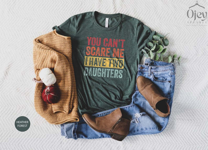 Puede incluir: Una camiseta verde con el texto "You can't scare me I have two daughters" en una fuente de estilo vintage. La camiseta est&aacute; colocada sobre una superficie blanca con un su&eacute;ter marr&oacute;n, jeans azules y botas marrones.