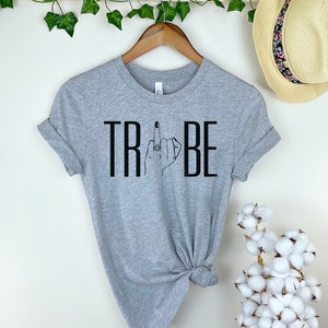 May include: Gray t-shirt with black text that reads "TRIBE" and a black line drawing of a hand giving the middle finger with a ring on the finger.