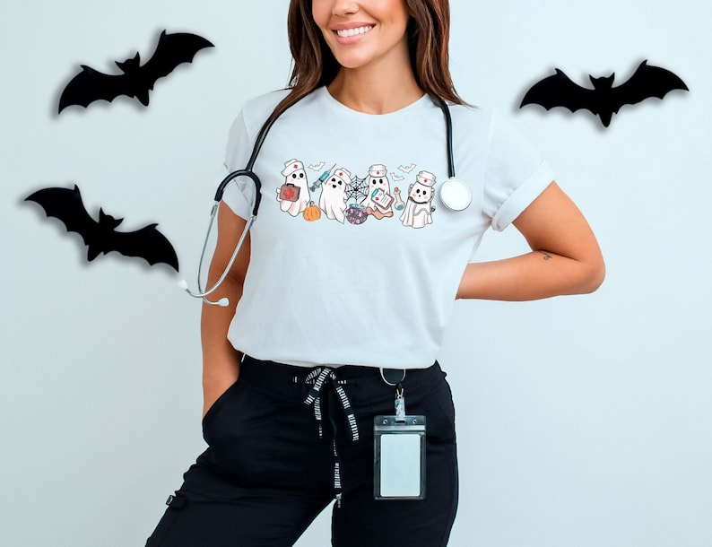 May include: A white t-shirt featuring a graphic of five ghosts dressed as nurses. The ghosts are holding medical supplies and a pumpkin. The t-shirt has a stethoscope around the neck.
