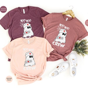 May include: Three t-shirts featuring a ghost wearing a nurse's hat and holding a bandage. The shirts are in heather maroon, heather mauve, and heather peach. The text on the shirts reads "Boo Boo Crew".
