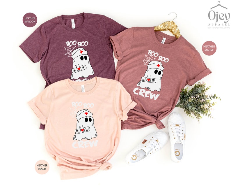 May include: Three t-shirts featuring a ghost wearing a nurse's hat and holding a bandage. The shirts are in heather maroon, heather mauve, and heather peach. The text on the shirts reads "Boo Boo Crew".