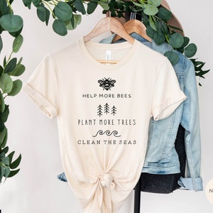 K&ouml;nnte beinhalten: Ein beiges T-Shirt mit einer schwarzen Bienengrafik und dem Text "Help More Bees, Plant More Trees, Clean the Seas". Das Shirt h&auml;ngt an einem Holzb&uuml;gel mit einer Jeansjacke und Gr&uuml;npflanzen im Hintergrund.