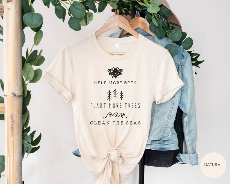 K&ouml;nnte beinhalten: Ein beiges T-Shirt mit einer schwarzen Bienengrafik und dem Text "Help More Bees, Plant More Trees, Clean the Seas". Das Shirt h&auml;ngt an einem Holzb&uuml;gel mit einer Jeansjacke und Gr&uuml;npflanzen im Hintergrund.