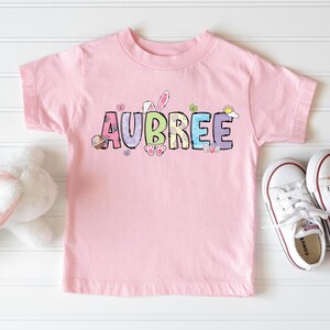 May include: A light pink t-shirt with the name "AUBREE" in colorful letters, decorated with Easter-themed graphics. White sneakers with red trim are next to the shirt. A white stuffed bunny is on the left.