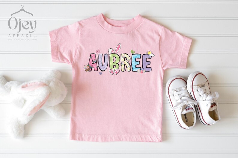 May include: A light pink t-shirt with the name "AUBREE" in colorful letters, decorated with Easter-themed graphics. White sneakers with red trim are next to the shirt. A white stuffed bunny is on the left.
