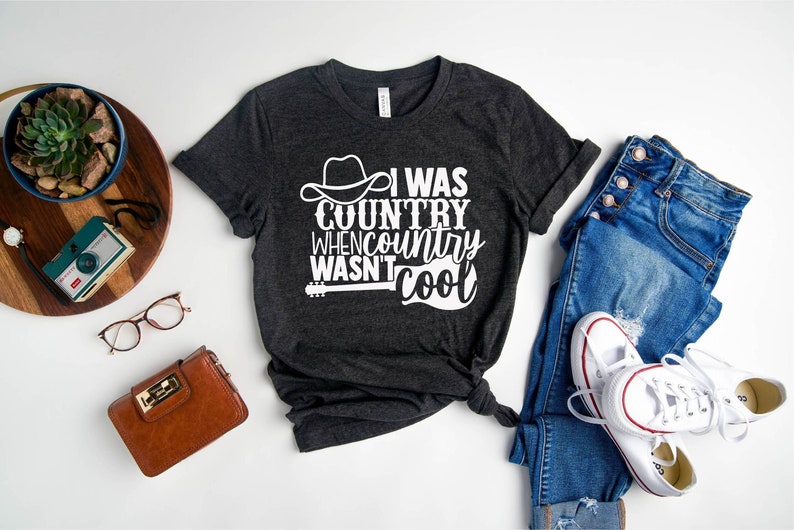 May include: A dark gray t-shirt with a white graphic that says "I was country when country wasn't cool". The graphic includes a cowboy hat and a guitar.