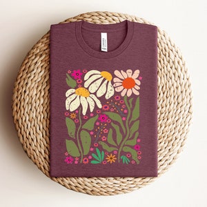 May include: A maroon t-shirt with a floral design featuring white daisies, pink flowers, and green leaves. The flowers are stylized and have a vintage feel.