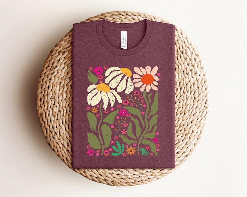 May include: A maroon t-shirt with a floral design featuring white daisies, pink flowers, and green leaves. The flowers are stylized and have a vintage feel.