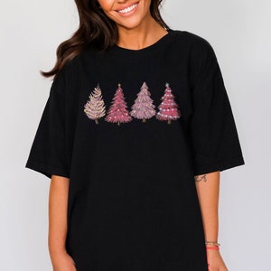May include: Black oversized t-shirt featuring a design of four pink Christmas trees with gold star and snowflake decorations.