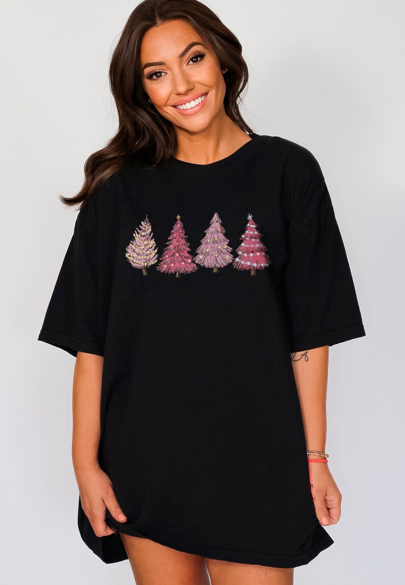 May include: Black oversized t-shirt featuring a design of four pink Christmas trees with gold star and snowflake decorations.