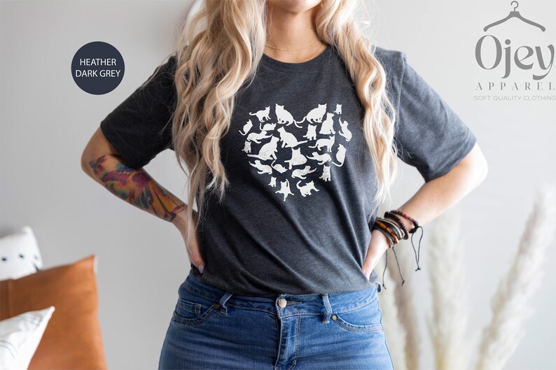 May include: A heather dark grey t-shirt with a white graphic of a heart made of cats. The text "Ojev Apparel Soft Quality Clothing" is visible in the upper right corner of the image.