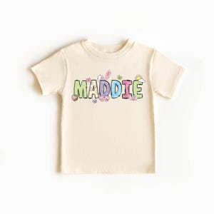 May include: Cream-colored short-sleeved t-shirt with the name "MADDIE" in colorful, patterned letters. Easter-themed graphics, including bunny ears, butterflies, and a sun, are incorporated into the design. A cute and festive children's tee.