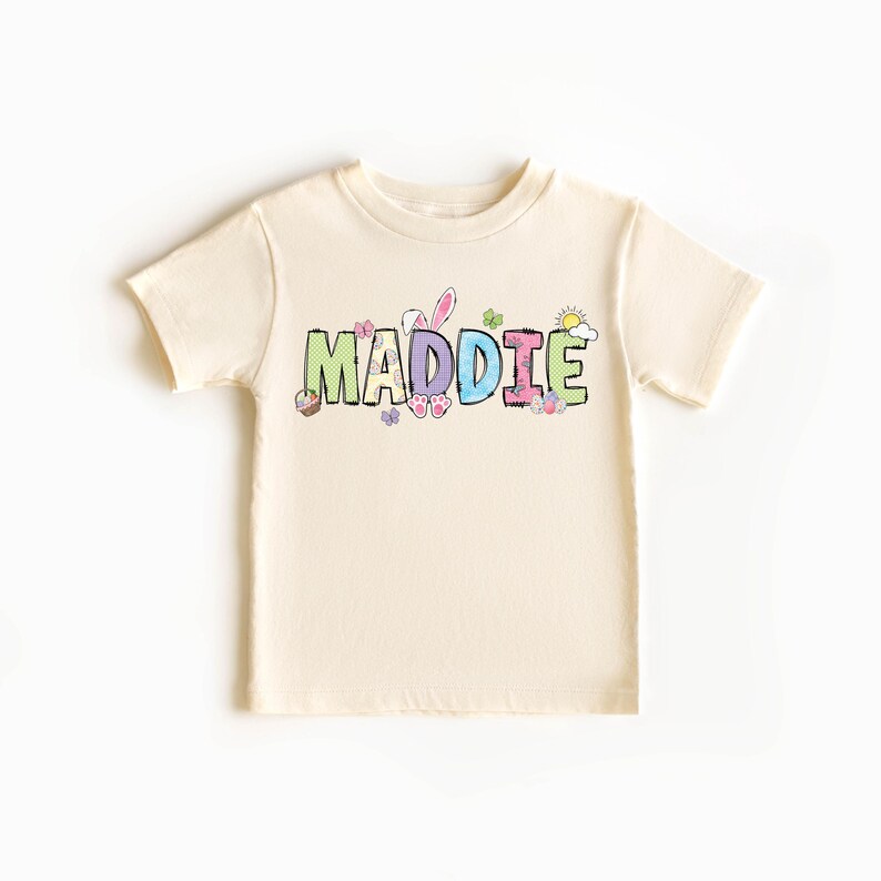 May include: Cream-colored short-sleeved t-shirt with the name "MADDIE" in colorful, patterned letters. Easter-themed graphics, including bunny ears, butterflies, and a sun, are incorporated into the design. A cute and festive children's tee.
