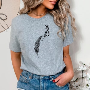 Op de afbeelding: Een grijs t-shirt met een zwarte afbeelding van een veer met vogels die eruit vliegen.