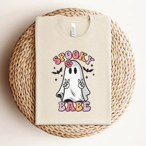 May include: A beige t-shirt with a graphic of a ghost wearing a flower crown and peace signs. The text "Spooky Babe" is printed below the ghost.