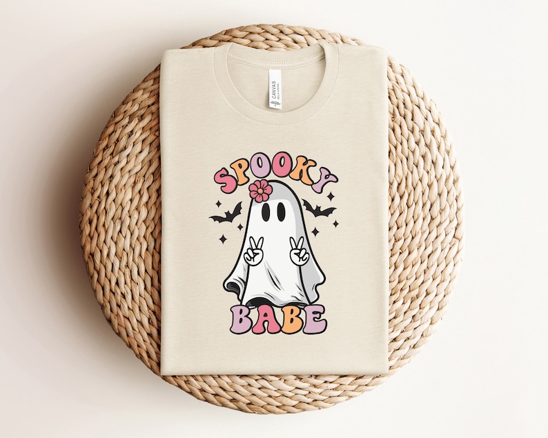 May include: A beige t-shirt with a graphic of a ghost wearing a flower crown and peace signs. The text "Spooky Babe" is printed below the ghost.