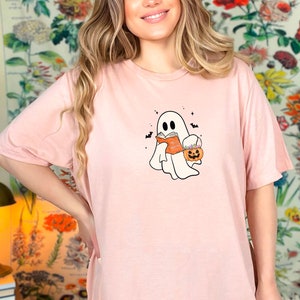 May include: A pink t-shirt with a white ghost holding a book and a pumpkin bucket. The ghost has a small smile and is surrounded by bats.