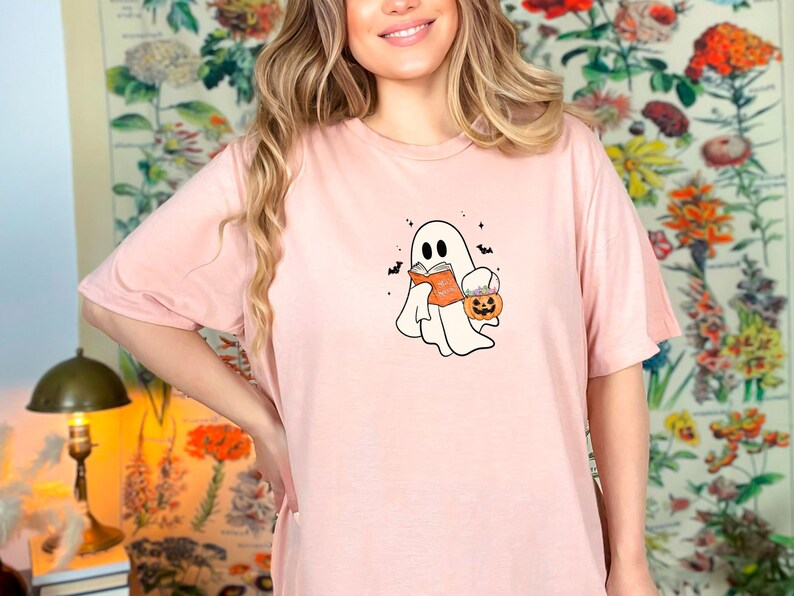 May include: A pink t-shirt with a white ghost holding a book and a pumpkin bucket. The ghost has a small smile and is surrounded by bats.