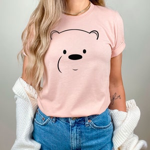 May include: A pink t-shirt with a black outline of a bear's face. The bear has a small black nose and two black eyes.