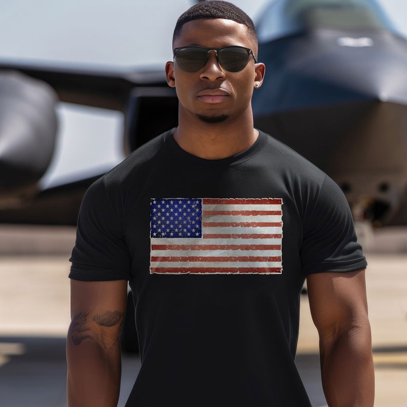 Pode incluir: Uma camiseta preta com um gr&aacute;fico de bandeira americana de estilo vintage impresso na frente. A bandeira &eacute; vermelha, branca e azul com um visual desgastado.