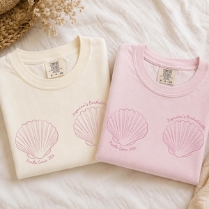 May include: Two crew neck t-shirts, one cream and one light pink, each featuring a dark pink line art graphic of two seashells. The text "Jasmine's Bachelorette" and "Punta Cana 2024" is printed in a cursive font near the seashell illustrations.