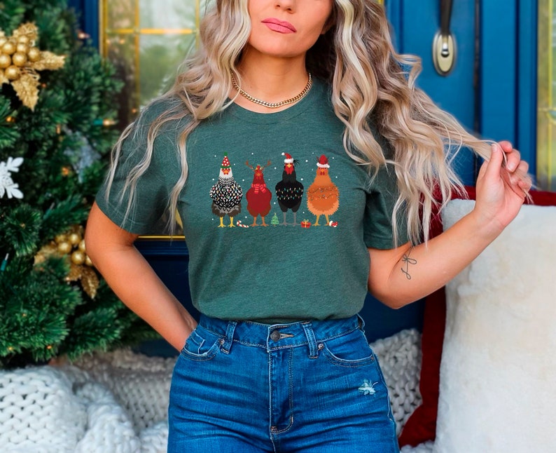 Może przedstawiać: Zielony T-shirt z świątecznym wzorem przedstawiającym cztery kury w czapkach Świętego Mikołaja trzymające świąteczne ozdoby. Kury stoją w rzędzie i patrzą w prz&oacute;d. T-shirt jest wykonane z miękkiego, wygodnego materiału i idealnie nadaje się na okres świąteczny.