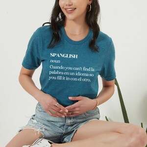Puede incluir: Una camiseta azul turquesa con la palabra "Spanglish" definida como "Cuando no encuentras la palabra en un idioma, la completas con el otro."