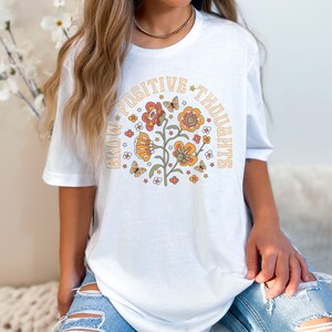 May include: White t-shirt with a graphic of a flower with butterflies and the text "Grow Positive Thoughts" in a retro style.
