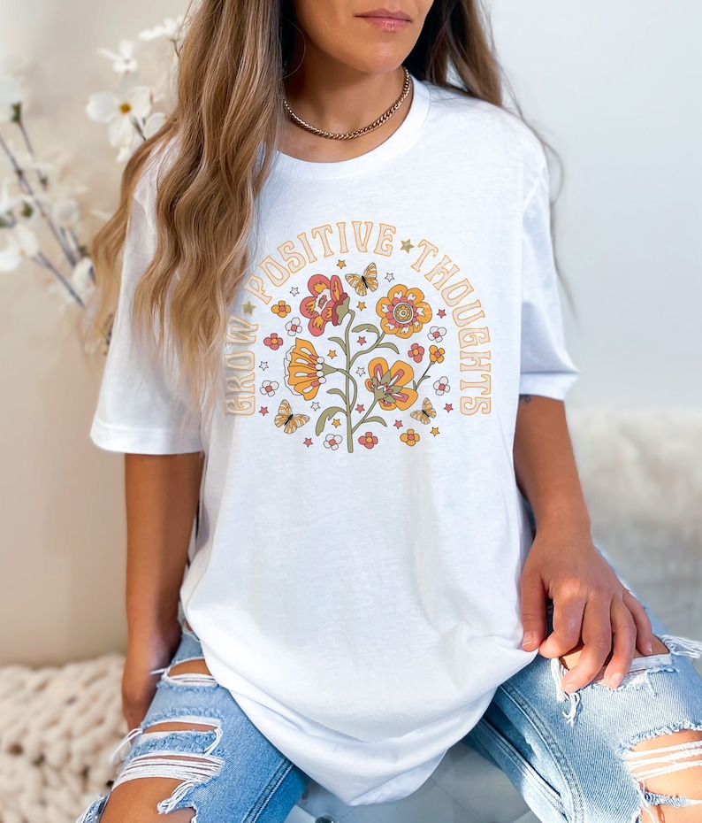 May include: White t-shirt with a graphic of a flower with butterflies and the text "Grow Positive Thoughts" in a retro style.