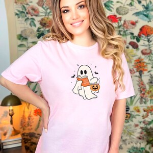 May include: A pink t-shirt with a white ghost holding a book that says "Boo Books" and a pumpkin bucket. The ghost has a cute face and is wearing a black headband.