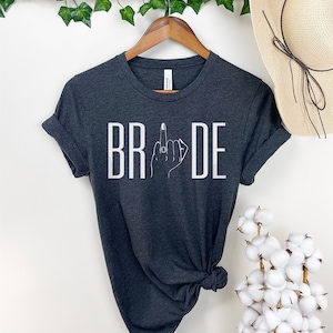 May include: A dark gray t-shirt with the word "BRIDE" printed in white. The letter "I" is replaced with a stylized image of a hand with a ring on the middle finger, giving a middle finger gesture.