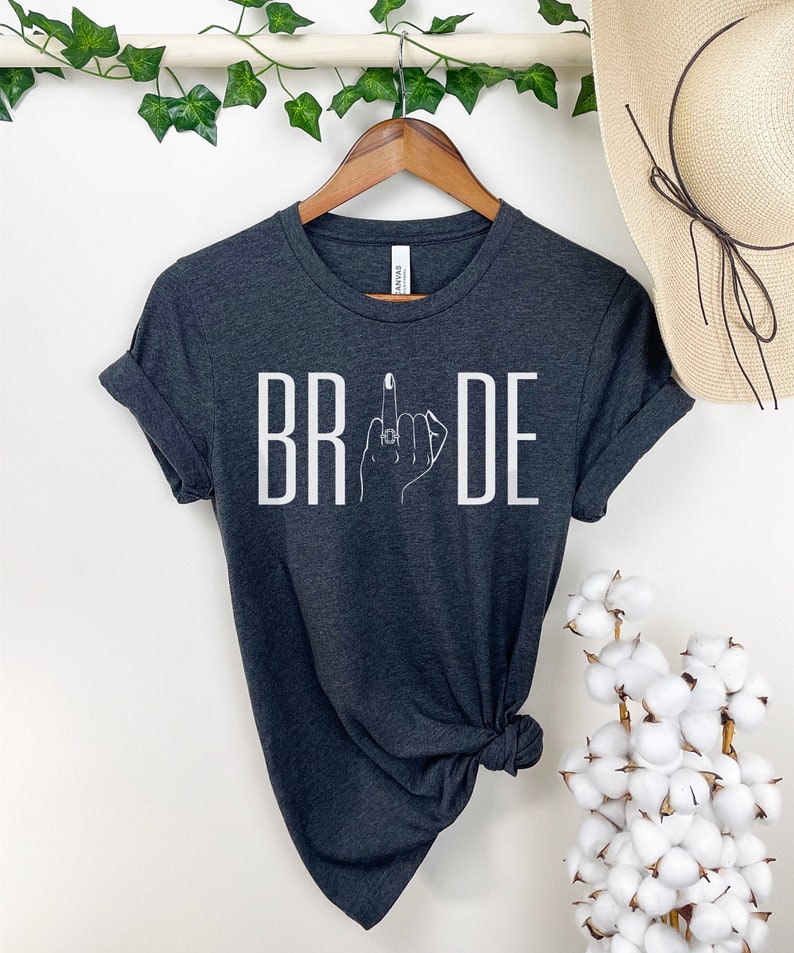 May include: A dark gray t-shirt with the word "BRIDE" printed in white. The letter "I" is replaced with a stylized image of a hand with a ring on the middle finger, giving a middle finger gesture.