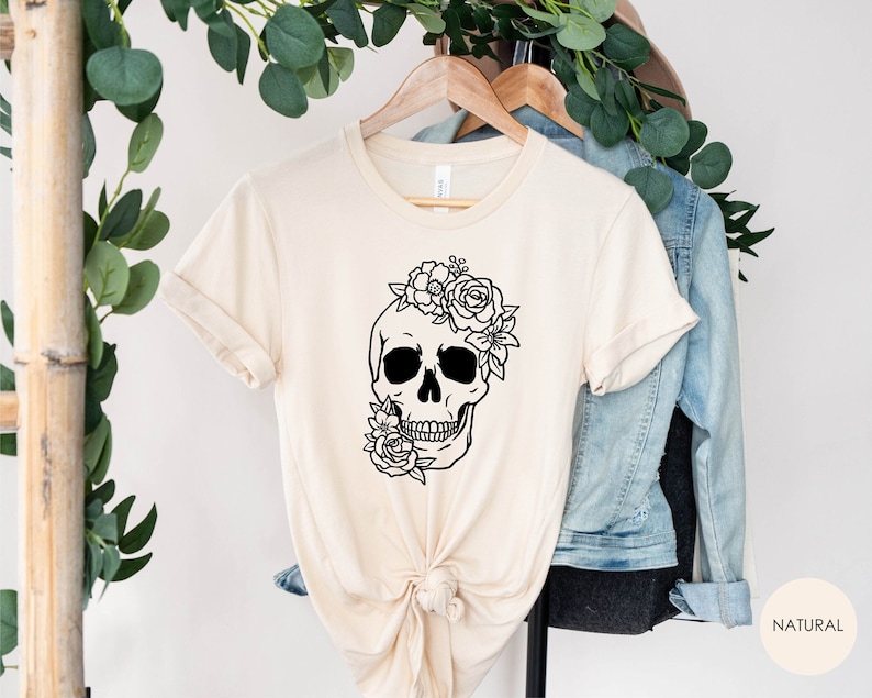 Peut inclure: Un t-shirt beige avec une illustration noir et blanc d'un cr&acirc;ne surmont&eacute; de fleurs. Le texte "NATURAL" est visible dans le coin inf&eacute;rieur droit de l'image.