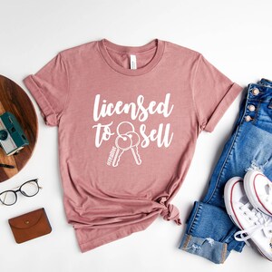 Pode incluir: Camiseta heather malva com o texto "Licensed to Sell" e um gr&aacute;fico de duas chaves. A camiseta est&aacute; disposta sobre uma superf&iacute;cie branca com um par de cal&ccedil;as de ganga, uma c&acirc;mara, um rel&oacute;gio e uns &oacute;culos.