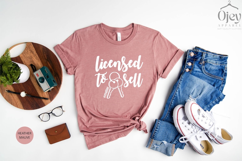 Pode incluir: Camiseta heather malva com o texto "Licensed to Sell" e um gr&aacute;fico de duas chaves. A camiseta est&aacute; disposta sobre uma superf&iacute;cie branca com um par de cal&ccedil;as de ganga, uma c&acirc;mara, um rel&oacute;gio e uns &oacute;culos.