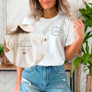 Peut inclure: Un t-shirt blanc avec un graphique sur le devant qui dit "Since 1776" avec un cercle d'&eacute;toiles autour du texte. Le dos du t-shirt a un graphique qui dit "The United States of America, The Land of the Free, The Home of the Brave" avec un petit drapeau am&eacute;ricain.