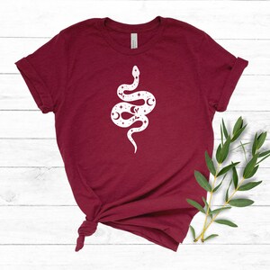 May include: A maroon t-shirt with a white graphic of a snake with stars and moons on its body.
