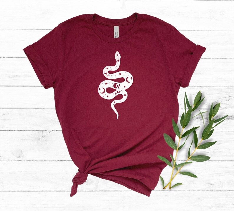 May include: A maroon t-shirt with a white graphic of a snake with stars and moons on its body.