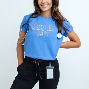 Puede incluir: Una mujer con una camiseta azul claro con el texto "MOTHER & BABY" en letras de colores. Tambi&eacute;n lleva pantalones negros y un estetoscopio alrededor del cuello.