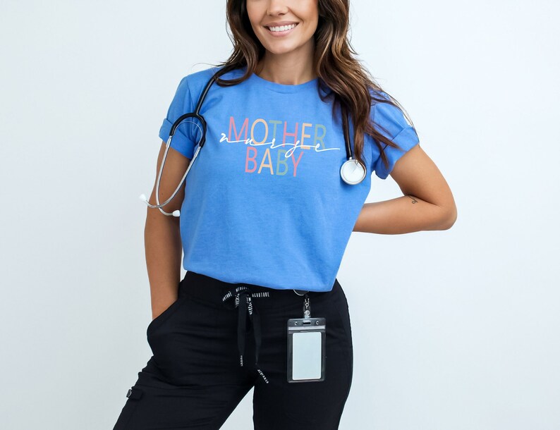 Puede incluir: Una mujer con una camiseta azul claro con el texto "MOTHER & BABY" en letras de colores. Tambi&eacute;n lleva pantalones negros y un estetoscopio alrededor del cuello.