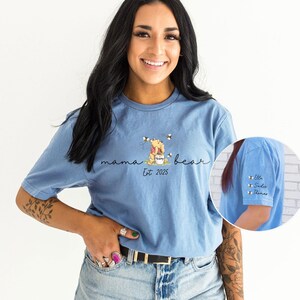 May include: A light blue t-shirt featuring a Winnie the Pooh illustration and the text "mama bear est 2025". The shirt has a small pocket on the sleeve with the names "Elle, Sadie, Thomas" written on it.