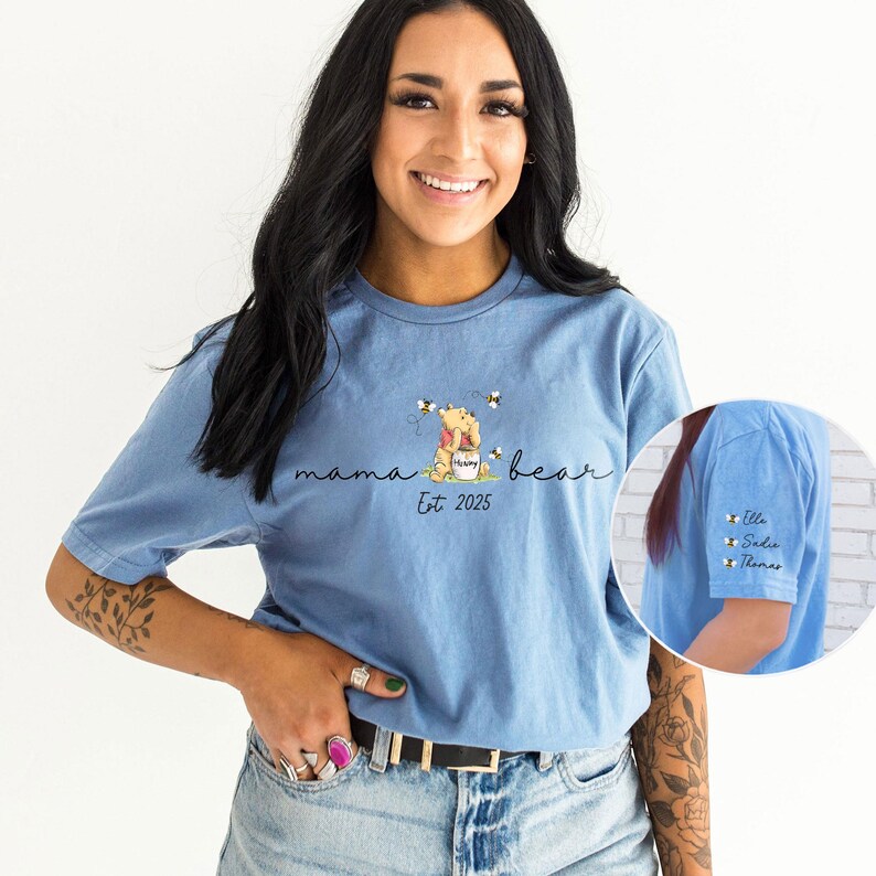 May include: A light blue t-shirt featuring a Winnie the Pooh illustration and the text "mama bear est 2025". The shirt has a small pocket on the sleeve with the names "Elle, Sadie, Thomas" written on it.