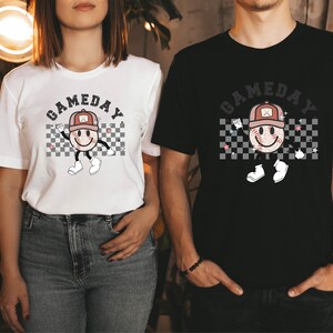 May include: Two people wearing white and black t-shirts with a graphic of a smiling baseball with a hat and the text "GAMEDAY" printed on the front.