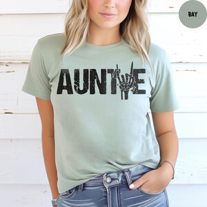 May include: A light green t-shirt with the word "AUNTIE" in distressed black lettering. The letter "E" is replaced with a hand gesture. The shirt is made of a soft material and is a casual style.