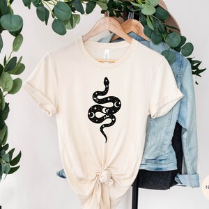May include: A beige t-shirt with a black snake design featuring moons and stars. The snake is curled up with its tail in its mouth. The t-shirt is hanging on a wooden hanger.