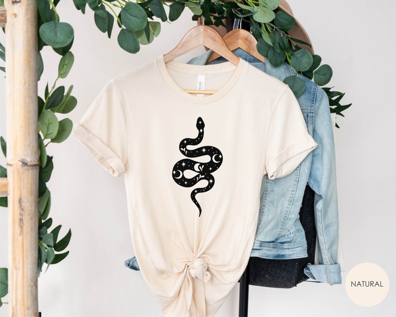 May include: A beige t-shirt with a black snake design featuring moons and stars. The snake is curled up with its tail in its mouth. The t-shirt is hanging on a wooden hanger.